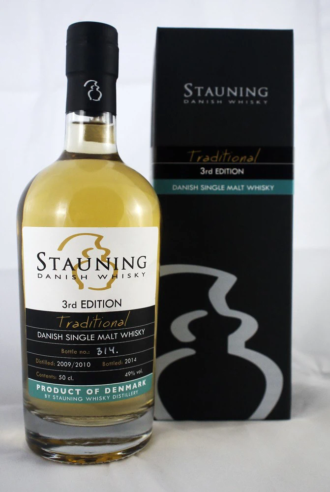 En flaske Stauning Danish Single Malt Whisky (3rd Edition) står ved siden af sin sorte og grå æske med guld- og blågrønne detaljer, begge med etiketten Traditional.